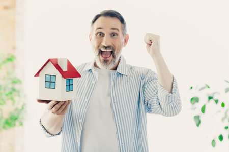 Cheerful mature man showcasing a model house with joy and enthusiasm in a brightly lit indoor...