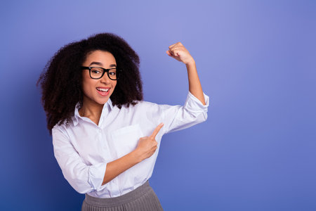 Confident young woman smiling and flexing her arm in empowering gesture on a purple backgroundの写真素材