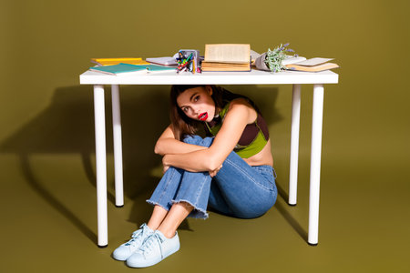 Young woman sitting under a white desk surrounded by books and stationary on a green background, wearing casual fashionの写真素材