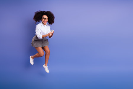 Young woman with curly hair jumps while holding a smartphone in her hands against a vibrant violet...