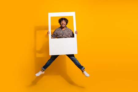 Energetic young man holding a photo frame jumping joyfully against a vibrant yellow background expressing positivityの写真素材