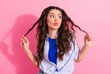 Young woman with wavy hair making funny gestures against a pink backdrop, showcasing playful attitude and casual styleの写真素材
