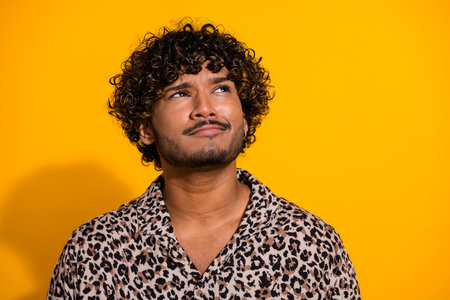 Young man with curly hair on a yellow background looking thoughtful wearing a stylish leopard print shirtの写真素材