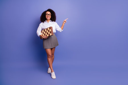 Confident young woman holding a chessboard while gesturing towards the side against a violet backgroundの写真素材
