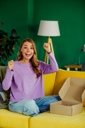 Young woman happily celebrating while sitting indoors with open cardboard box in cheerful home...