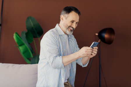 Mature businessman enjoying leisure time using smartphone in stylish modern home interior with green...