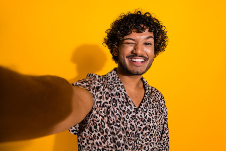 Cheerful young man takes a selfie against bright yellow background with charming smile wearing fashionable leopard print shirtの写真素材