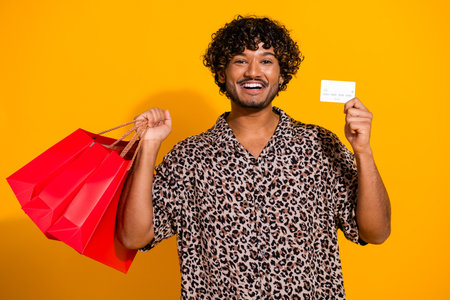 Funny man smiling confidently while holding vibrant shopping bags and credit card on a bold yellow...