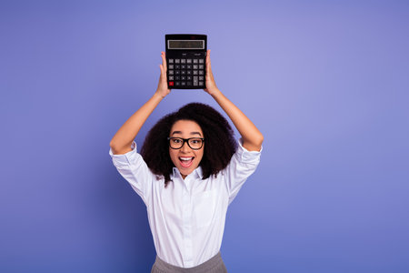 Lovely businesswoman holding calculator in joyful pose against purple background, showcasing success and financial expertiseの写真素材