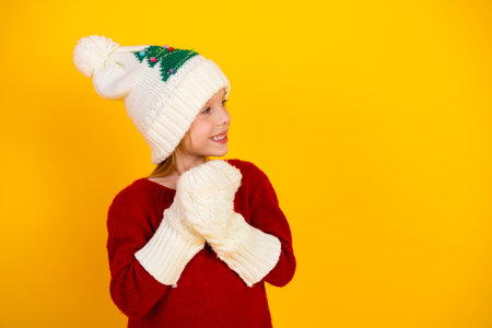 Joyful kid wearing red sweater and Christmas knitwear poses happily with festive hat and mittens against bright yellow background perfect for holiday season imageryの写真素材