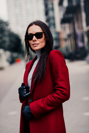 Portrait of a fashionable woman with brunette hair in a red coat and sunglass outdoors on a stylish urban streetの写真素材