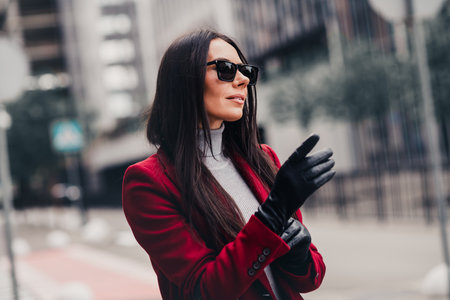 Stylish businesswoman in a red coat enjoying a sunny day outdoors in an urban setting embodying elegance and charmの写真素材