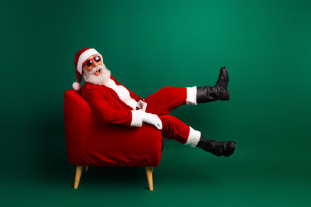 Santa Claus in a red armchair with sunglass sits stylishly against a vibrant green background celebrating Christmas joyの写真素材