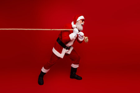Festive Santa Claus pulling a rope with strength against a red background, demonstrating holiday cheer and winter traditionsの写真素材