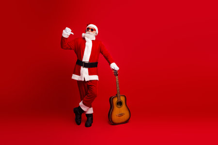 Santa Claus in a festive hat holding a guitar, posing on a red background with confidence and Christmas cheerの写真素材