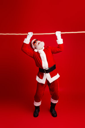 Santa Claus performing fun exercise poses with rope on vibrant red background symbolizing holiday spirit and fitnessの写真素材
