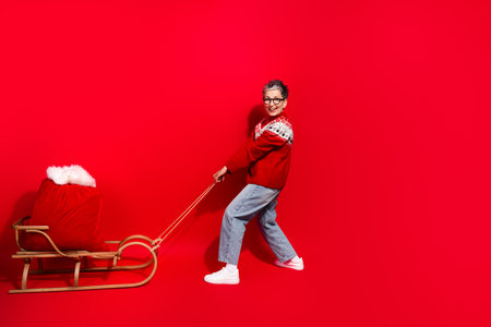 Festive elderly woman wearing a Christmas sweater in a red background pulling a sled filled with presents joyfullyの写真素材