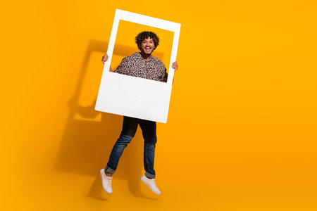 Funny man holding a frame while jumping joyfully against a bright yellow background wearing a leopard print shirtの写真素材