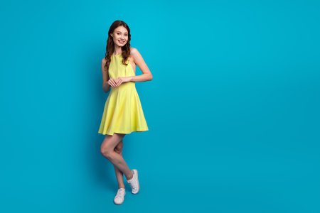 Stylish brunette woman in yellow dress posing confidently against a vibrant blue background, showcasing youthful fashion beautyの写真素材