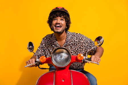 Cheerful young man with a trendy style riding a red scooter in front of a vibrant yellow backgroundの写真素材