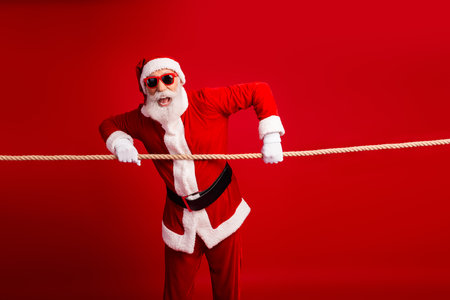Joyful Santa Claus in festive attire posing with rope prop on a vibrant red background during the Christmas seasonの写真素材