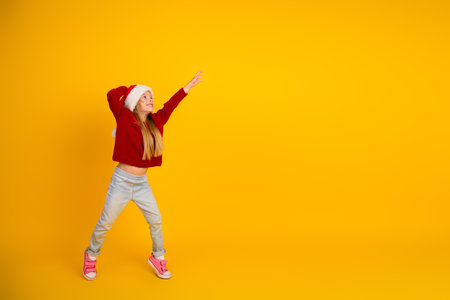 Funny cheerful girl in santa hat dances in bright yellow studio wearing red sweater and jeans celebrating christmas season joy and playful energyの写真素材
