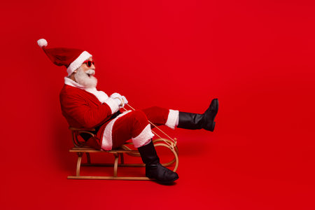 Santa Claus in a festive red costume sitting on a wooden sled wearing sunglass against a vibrant red backgroundの写真素材