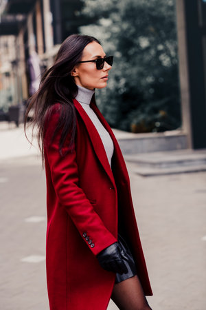 Stylish brunette woman wearing a red coat and sunglass walking outdoors in an urban setting on a sunny autumn dayの写真素材