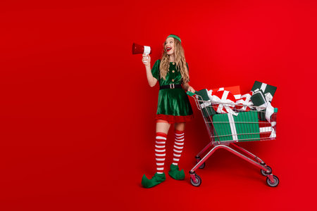 Festive elf showcasing holiday gifts with a megaphone and joyous cheer, surrounded by colorful packages on a vibrant red backdropの写真素材