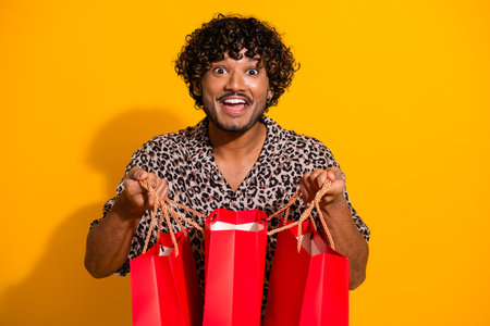 Excited young man enjoying a shopping spree holding red bags on a vivid yellow background depicting fun and styleの写真素材