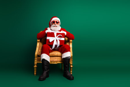 Santa Claus in a trendy costume sitting on a golden chair holding a gift box with a green background, Christmas cheerの写真素材