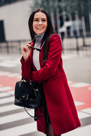 Beautiful businesswoman wearing elegant red coat outdoors in an urban street setting during fall or springの写真素材