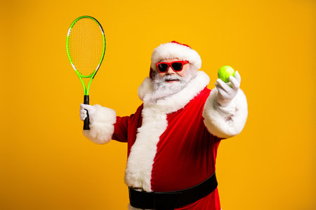 Santa Claus in festive attire holds a tennis racket and ball with a cheerful smile against a vibrant yellow backgroundの写真素材