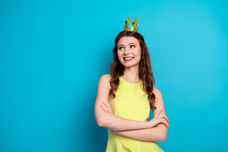 Happy young woman wearing a yellow dress and crown standing confidently against a vibrant blue backgroundの写真素材