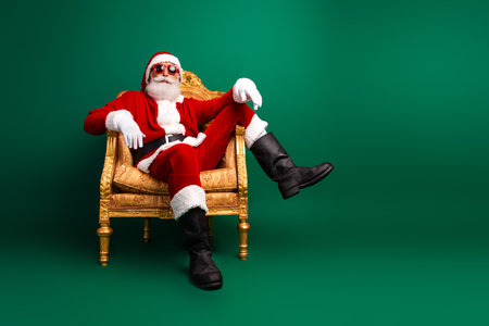 Stylish Santa with sunglass relaxing on a gold chair against a vibrant green background celebrating Christmas in styleの写真素材