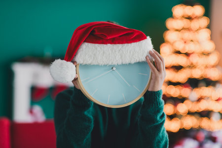 A whimsical festive scene showcasing a clock wearing a Santa hat surrounded by cozy Christmas decor and a glowing treeの写真素材