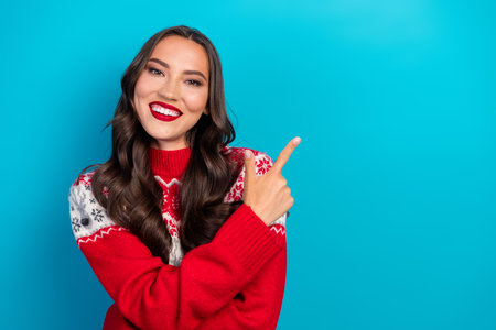 Festive young woman in red sweater smiling joyfully while pointing to highlight the holiday spirit on blue backgroundの写真素材