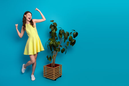 Cheerful young woman in a yellow dress posing next to a potted plant against a vibrant blue backgroundの写真素材