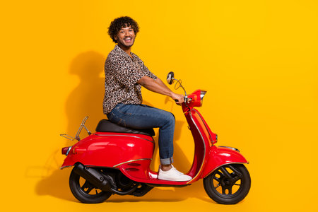 Young man riding a red scooter against yellow background wearing casual leopard print shirt, showcasing stylish leisure vibesの写真素材