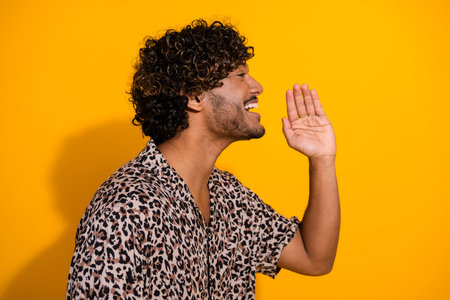 Young man in leopard print shirt gesturing loudly with hand near mouth on vibrant yellow backgroundの写真素材