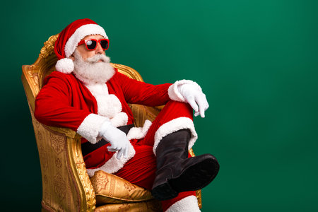 Stylish Santa Claus in a festive Christmas costume relaxing with sunglass on a luxurious chair against a green backgroundの写真素材