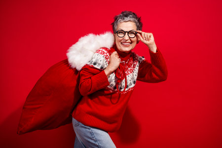 Festive senior woman in red with Santa bag celebrates Christmas joyfully against vibrant backgroundの写真素材