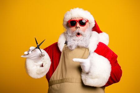 Santa Claus in funky red sunglasses holding scissors and pointing, festive holiday scene on yellow studio backgroundの写真素材