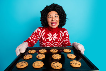 Joyful woman holding a tray of fresh cookies wearing a festive red sweater against a cheerful blue backgroundの写真素材