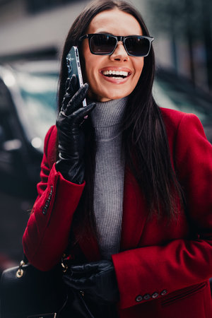 Confident businesswoman in a red coat outdoors talking on smartphone, enjoying urban lifestyle in stylish high fashion.の写真素材