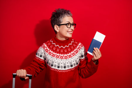Festive woman in cozy Christmas sweater holding passport and preparing for a holiday getaway against a red backgroundの写真素材