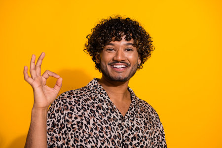 Stylish young man in leopard print shirt showing OK gesture against bright yellow background with cheerful expressionの写真素材