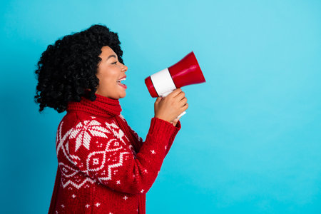Joyful young woman in festive red sweater with megaphone on vibrant blue backgroundの写真素材