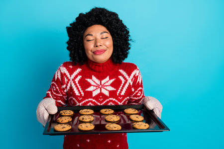 Young woman wearing festive sweater holding fresh baked cookies, happily celebrating the holiday season with a cheerful smile.の写真素材