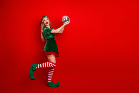 Festive woman dressed as Christmas elf holding a shiny ornament on a vibrant red background in a cheerful poseの写真素材